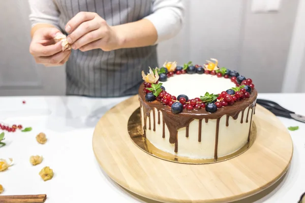 Il pasticcere decora con bacche una torta di biscotto con crema bianca e cioccolato. Cake si trova su un supporto di legno su un tavolo bianco. Il concetto di pasticceria fatta in casa, torte di cottura . — Foto Stock