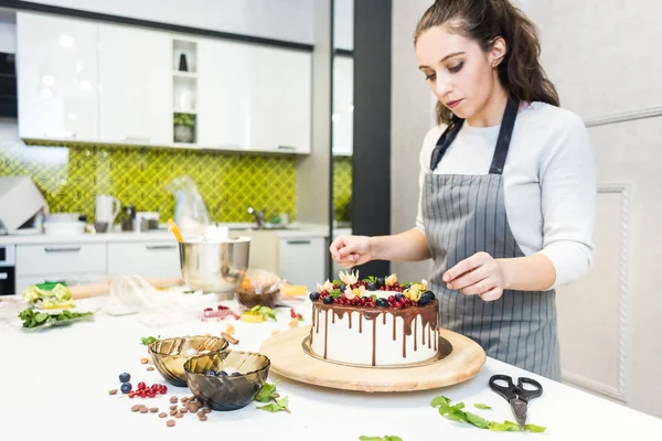 Cukrászda díszíti bogyókat keksz torta, fehér tejszín és csokoládé. A sütemény egy fából készült állványon áll egy fehér asztalon. A koncepció a házi készítésű péksütemények, főzősütemények. — Stock Fotó