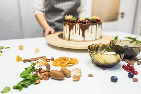 De banketbakkerij versiert met bessen een koekjes cake met witte room en chocolade. Cake staat op een houten stand op een witte tafel. Het concept van zelfgemaakte gebak, koken cakes. — Stockfoto