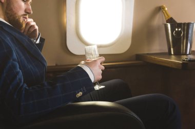 Pahalı bir takım elbiseli Genç başarılı bir işadamı elinde bir bardak şampanya ile özel bir Jet sandalyesinde oturur ve pencereden dışarı bakıyor