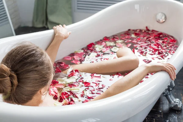 Молода приваблива дівчина приймає ванну кімнату з пелюстками квітів і відпочиває на тлі красивого світлого інтер'єру. Спа-процедури для краси та здоров'я при догляді за шкірою — стокове фото