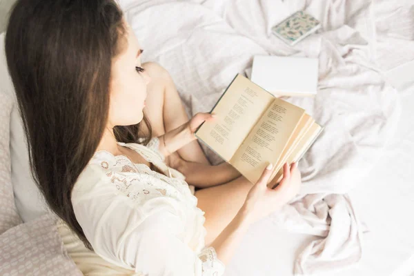 Aantrekkelijk jong meisje in een nightgown zit in bed en leest een boek — Stockfoto