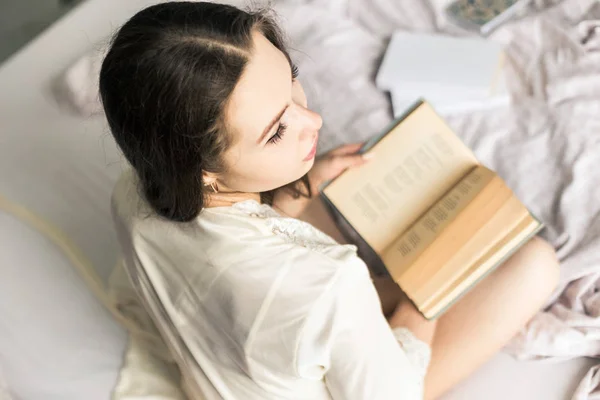 Atractiva joven en un camisón se sienta en la cama y lee un libro —  Fotos de Stock