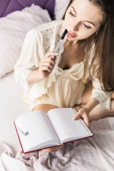 Aantrekkelijk jong meisje in een nachtjapon zit in bed en schrijft in een notitieboekje. — Stockfoto