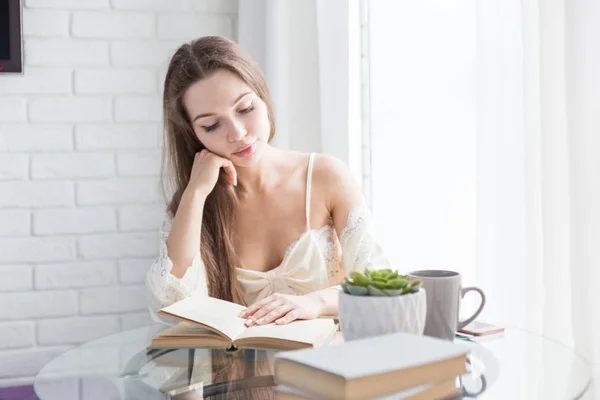 Mooi jong meisje in een nightgown zit in de ochtend aan de glazen tafel, kijkt uit het raam en leest een boek — Stockfoto