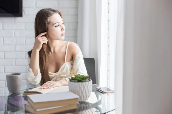 Menina bonita em uma camisola senta-se de manhã na mesa de vidro, olha pela janela e lê um livro — Fotografia de Stock