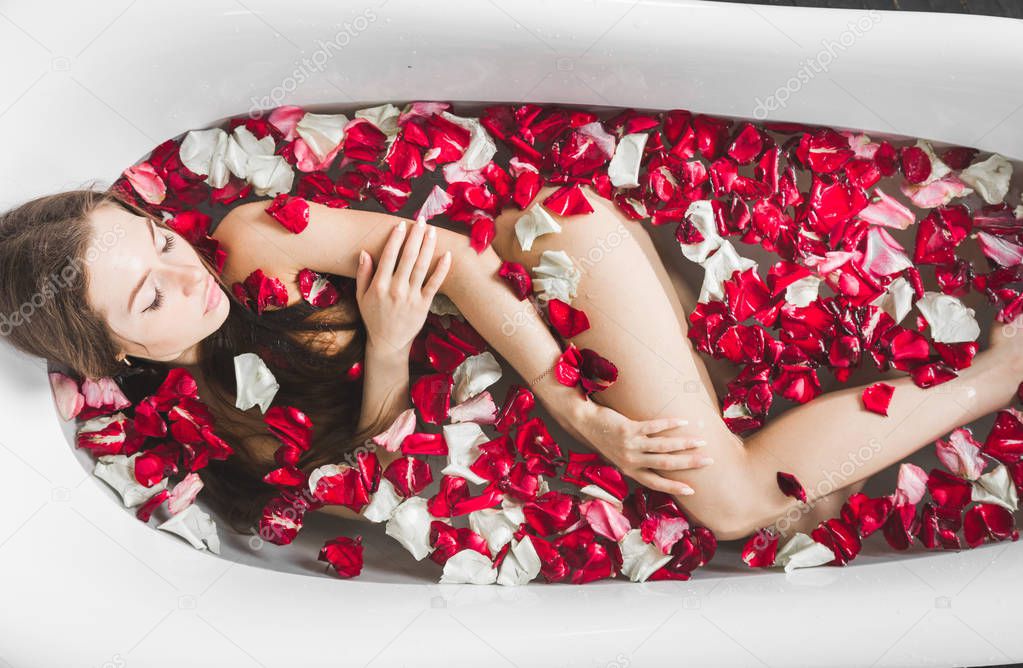 A young attractive girl takes a bathroom with flower petals and relaxes against the background of a beautiful light interior. Spa treatments for beauty and health with skin care