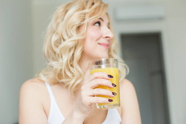 Attractive blonde woman sits on the edge of the bed at home and holds a stocker with orange juice in her hand. Morning mood and vigor