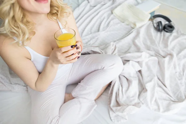 Приваблива блондинка сидить на краю ліжка вдома і тримає в руці акціонер з апельсиновим соком. Ранковий настрій і енергійність — стокове фото