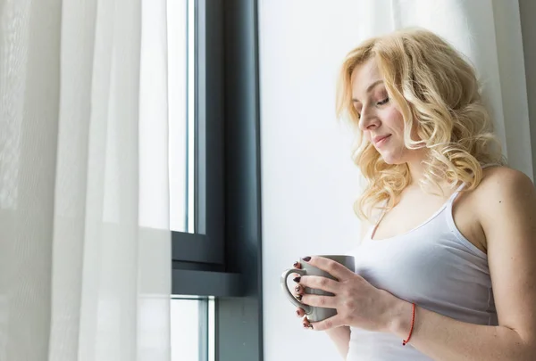 Aantrekkelijke blonde vrouw staat bij het raam met een kopje koffie of thee — Stockfoto