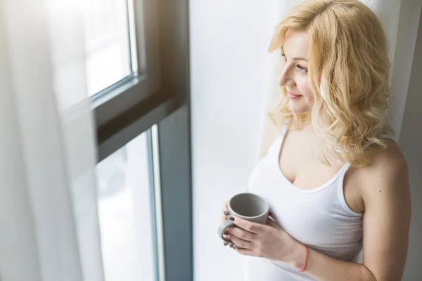 Portret van een aantrekkelijke blonde vrouw die bij het raam staat met een kop koffie of thee — Stockfoto