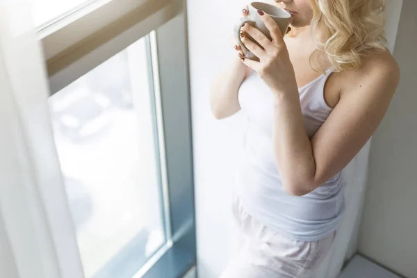 Attractive blonde woman stands near the window with a cup of coffee or tea — Stock Photo, Image