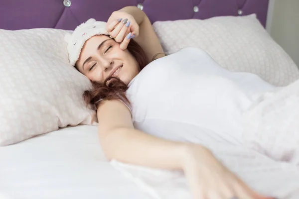 Een aantrekkelijk jong brunette meisje vrouw wakker in haar bed in een slaap masker. — Stockfoto