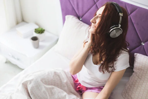 Atractiva joven morena se sienta por la mañana en su cama con auriculares y un teléfono inteligente y escucha música de buen humor — Foto de Stock