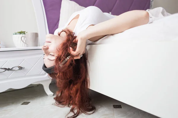 En attraktiv ung brunett kvinna ligger i sin säng på morgonen med huvudet hängande från kanten med hörlurar och en smartphone och lyssna på musik på ett gott humör — Stockfoto