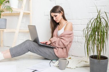 Genç ve mutlu bir kadın parlak bir apartman dairesinde ya da ofis içinde yerde oturuyor ve bir dizüstü bilgisayarda çalışıyor, iş yerinde serbest çalışan bir kız, uzaktan öğrenme öğrencileri, evrak işleri ve çevrimiçi öğrenim kavramı