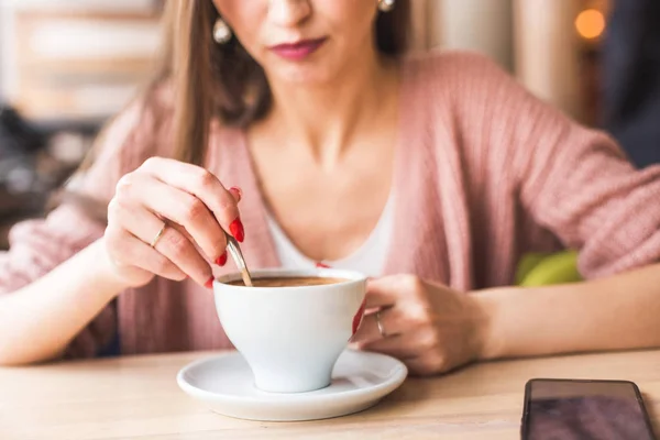 Jovem atraente senta-se a uma mesa em um café com uma xícara de café e mistura com uma colher — Fotografia de Stock