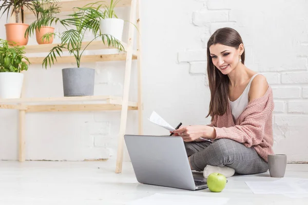 Mujer joven se sienta en el suelo en un interior de apartamento escandinavo con una computadora portátil, estudiar derecho, chica independiente en el trabajo, estudiante de educación a distancia, empleo en línea, papeleo y concepto de externalización — Foto de Stock