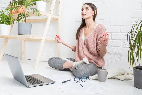 Молода щаслива жінка сидить на підлозі в позі йоги в світлій квартирі і працює за ноутбуком, дівчина-фрілансер медитує під час роботи. робота під напругою — стокове фото