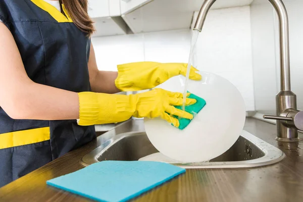 Une jeune femme en gants jaunes lave la vaisselle avec une éponge dans l'évier. Maison service de nettoyage professionnel. — Photo