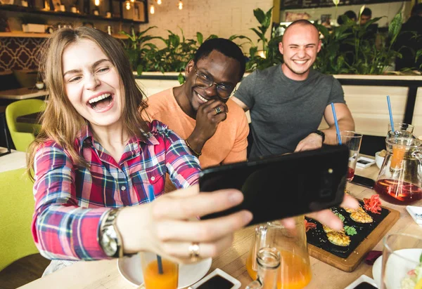 Група молодих веселих друзів сидить у кафе, їсть, п'є напої. Друзі беруть селфі і фотографують . — стокове фото