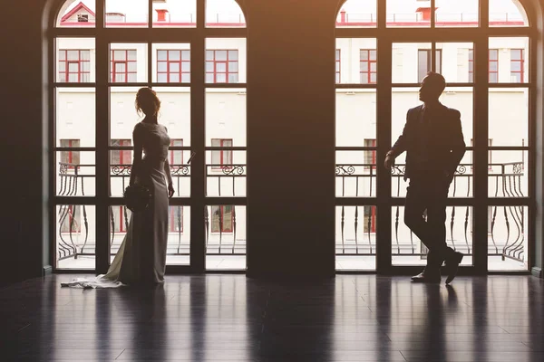 light shadow silhouettes of a man in a suit and a woman in a dress and with a bouquet of flowers facing each other near a large panoramic stained-glass window in a chic empty interior