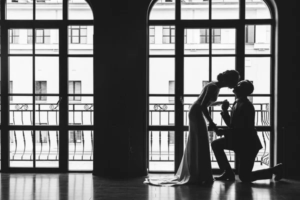 Silhouettes of a man in a suit and women in a dress and with a bouquet of flowers. The man knelt down in front of his lover, took her hand and kissed her near a large panoramic stained glass window.