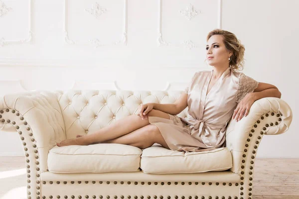 Hermosa mujer rubia sexy joven en una bata de seda sentada en un sofá — Foto de Stock