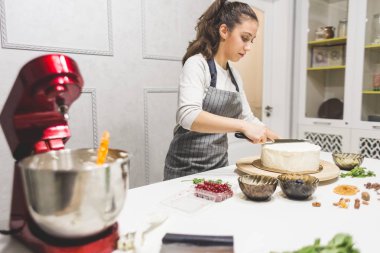Ön planda bir krema ve kırmızı elektrikli mikser ile bir metal kase. Arka planda, şekerleştirici bir bisküvi pastası üzerinde beyaz krem yumuşatın bir mutfak spatula ile.