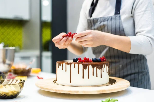 De banketbakkerij versiert met bessen een koekjes cake met witte room en chocolade. Cake staat op een houten stand op een witte tafel. Het concept van zelfgemaakte gebak, koken cakes. — Stockfoto