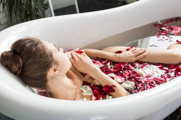 Una chica joven y atractiva toma un baño con pétalos de flores y se relaja sobre el fondo de un hermoso interior de luz. Tratamientos de spa para la belleza y la salud con cuidado de la piel — Foto de Stock