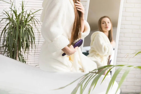Attraente ragazza in un accappatoio bianco pettinando i capelli in piedi davanti allo specchio all'interno del bagno e facendo procedure mattutine. Bellezza e cura del corpo — Foto Stock