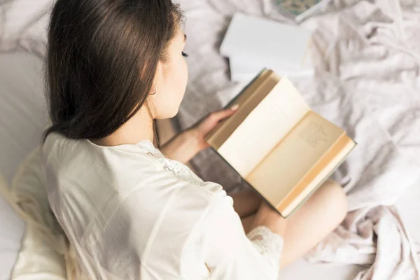 Aantrekkelijk jong meisje in een nightgown zit in bed en leest een boek — Stockfoto