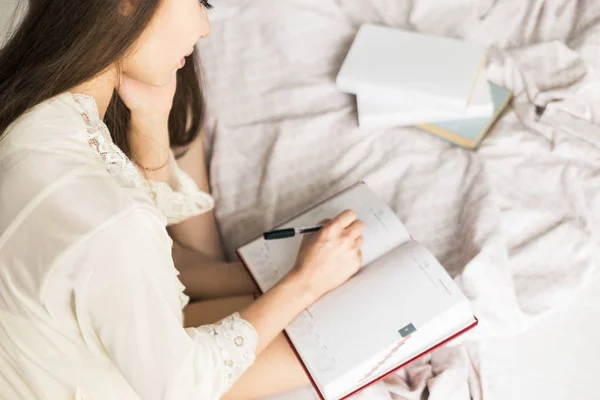 Aantrekkelijk jong meisje in een nachtjapon zit in bed en schrijft in een notitieboekje. — Stockfoto