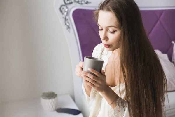 Menina atraente em roupa de noite senta-se na borda da cama junto à janela com uma xícara de café — Fotografia de Stock