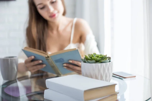 Hermosa joven en un camisón se sienta por la mañana en la mesa de cristal, mira por la ventana y lee un libro — Foto de Stock