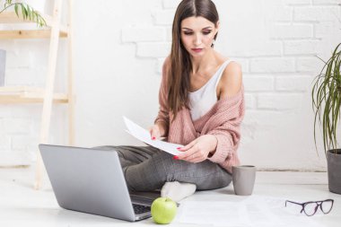 Genç ve mutlu bir kadın parlak bir apartman dairesinde ya da ofis içinde yerde oturuyor ve bir dizüstü bilgisayarda çalışıyor, iş yerinde serbest çalışan bir kız, uzaktan öğrenme öğrencileri, evrak işleri ve çevrimiçi öğrenim kavramı