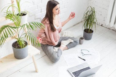 Genç, mutlu bir kadın yoga pozunda, parlak bir dairede oturuyor ve dizüstü bilgisayarın arkasında çalışıyor. Serbest çalışan bir kız çalışırken meditasyon yapıyor. İş stresi.