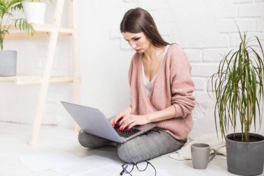 Genç ve mutlu bir kadın parlak bir apartman dairesinde ya da ofis içinde yerde oturuyor ve bir dizüstü bilgisayarda çalışıyor, iş yerinde serbest çalışan bir kız, uzaktan öğrenme öğrencileri, evrak işleri ve çevrimiçi öğrenim kavramı