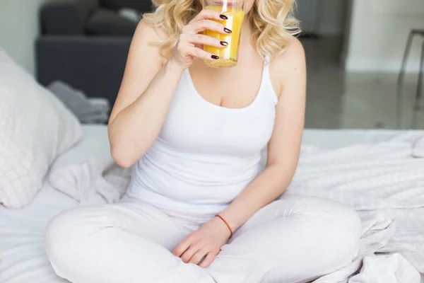 魅力的なブロンドの女性は自宅でベッドの端に座っており、彼女の手にオレンジジュースとストックを保持しています。朝の気分と活力 — ストック写真