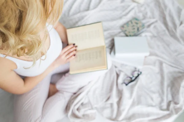 Aantrekkelijke blonde vrouw in bril met zwarte rand zit aan de rand van het bed thuis met een boek en leest — Stockfoto