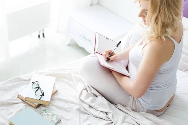 Aantrekkelijke blonde vrouw zit aan de rand van het bed thuis en schrijft in een notitieboekje — Stockfoto