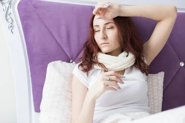 Een aantrekkelijke jonge brunette zit in haar bed met een thermometer. Verkoudheid en thuisbehandeling — Stockfoto