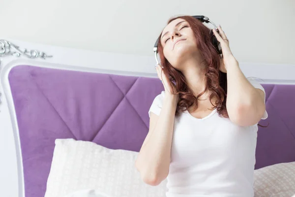 Jolie jeune femme brune s'assoit le matin dans son lit avec un casque et un smartphone et écoute de la musique de bonne humeur — Photo