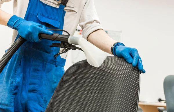 Jovem em vestuário de trabalho e luvas de borracha limpa a cadeira de escritório com equipamento profissional . — Fotografia de Stock