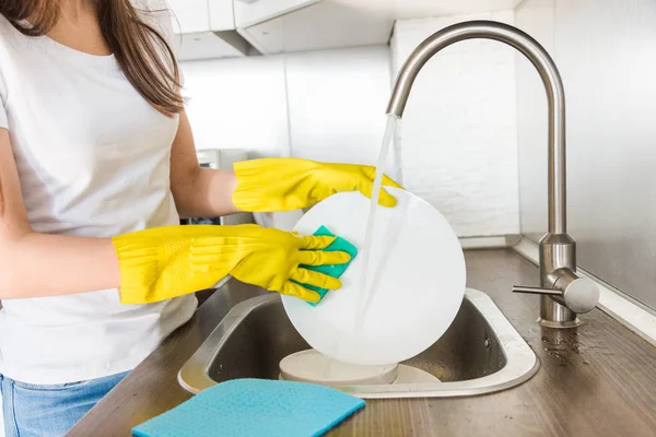 Eine junge Frau in gelben Handschuhen spült Geschirr mit einem Schwamm in der Spüle. Professioneller Reinigungsservice. — Stockfoto