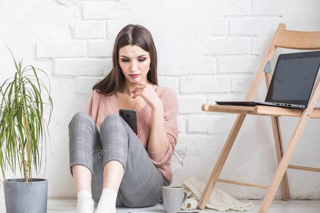 A happy woman in homemade casual clothes sits in a bright interior and uses a telephone, the girl is resting while working at a laptop and surfing the Internet. Cozy freelance work space,