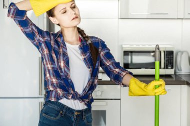Genç bir kadın evde temizlik yapar, mutfağı yıkar. Masada paçavralar ve sarı eldivenlerle dolu bir kova.