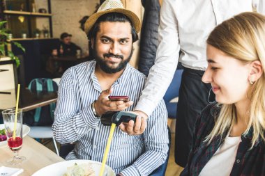 Genç, çekici Hindu adam, kafeteryada, bağlantısız akıllı telefon ödemesiyle ödeme yapıyor.