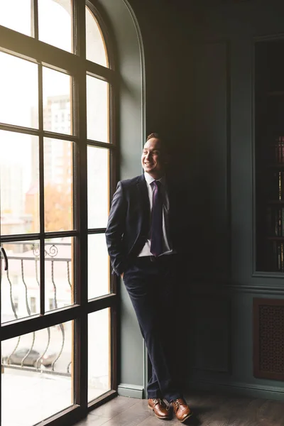 Retrato de un exitoso hombre de negocios de pie en un costoso traje de negocios cerca de la ventana de su oficina y sonriendo — Foto de Stock